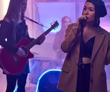 A person standing with a mic and another person with a guitar standing behind them